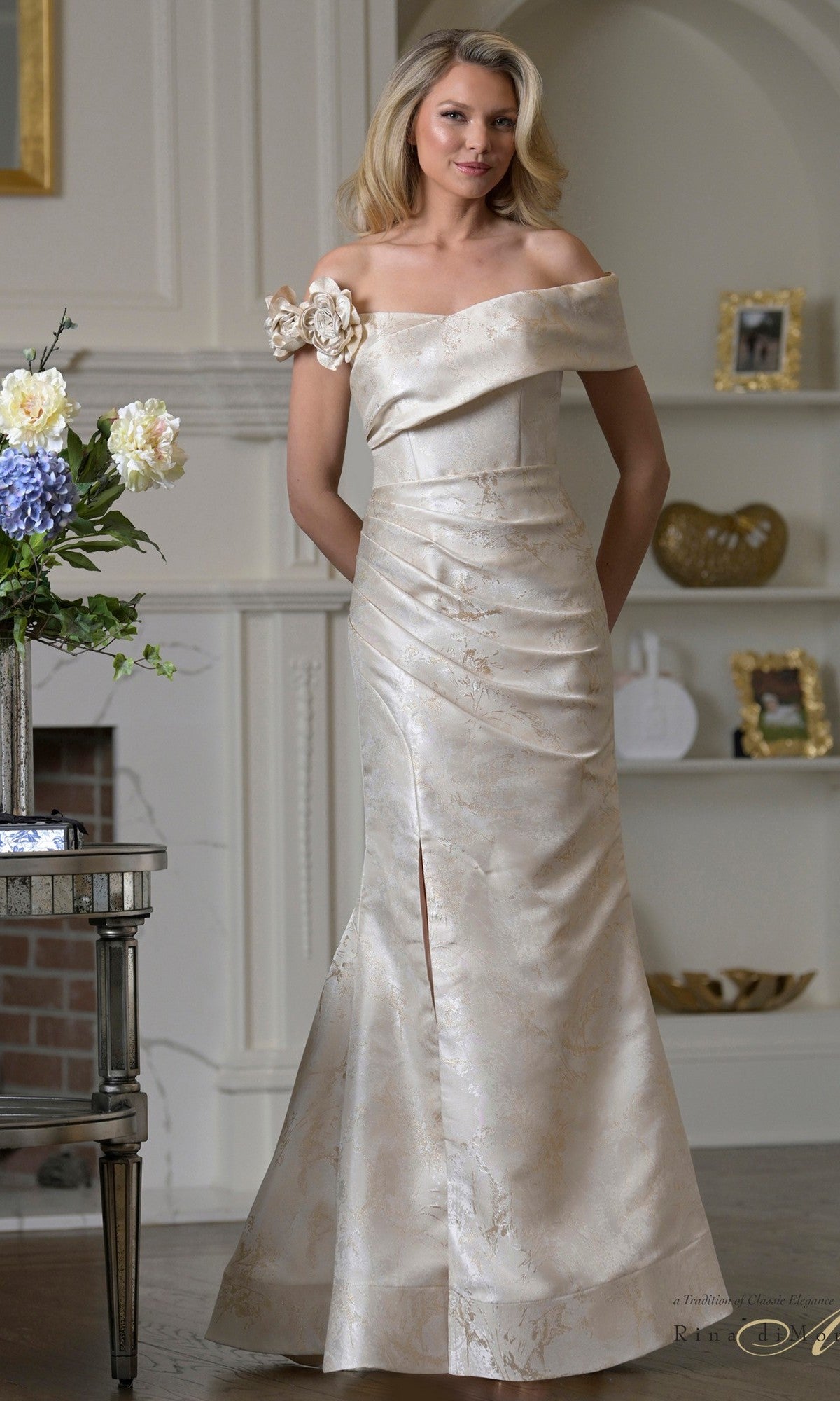 A woman stands indoors wearing the Rina Di Montella RD3522 long formal dress in champagne satin, featuring floral embellishment on one shoulder and elegant off-shoulder sleeves—perfect for any formal occasion.